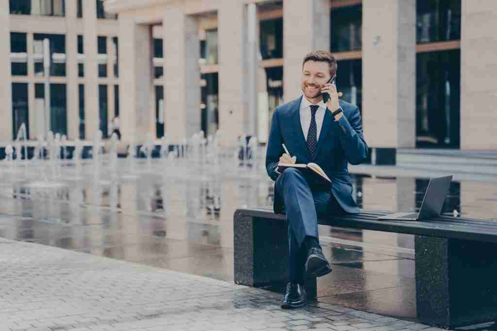 Successful businessman talking on mobile phone while working outside, sitting on bench